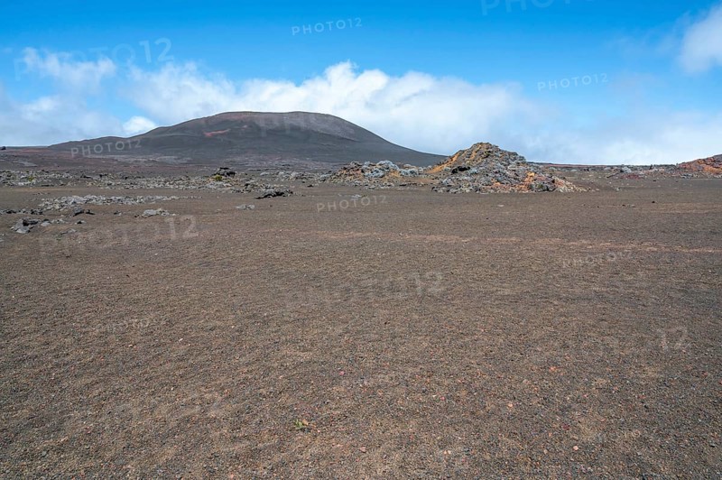 The Route du Volcan crosses the vast, rust-red volcanic desert of the Plaine des Sables, whose surface is dominated at extreme close angles by lava granules and sparse, barren grasses, while in the background a black-brown volcanic cone rises steeply into the blue sky under white cloud banks, Plaine des Sables, Highlands, La Reunion, France
