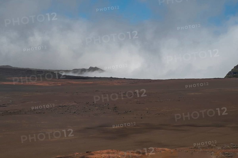 The Route du Volcan leads through the vast, rust-red lunar landscape of the Plaine des Sables, which is interspersed with dark lava rocks, while clouds hang over the eroded, steep volcanic rocks on the horizon, Plaine des Sables, Highlands, La Reunion, France
