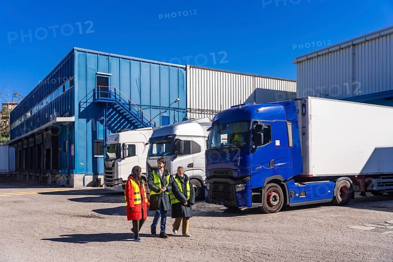 Logistics team in high visibility vests walking and talking beside refrigerated semitrailers at a cold storage facility on a sunny day, coordinating transport and loading