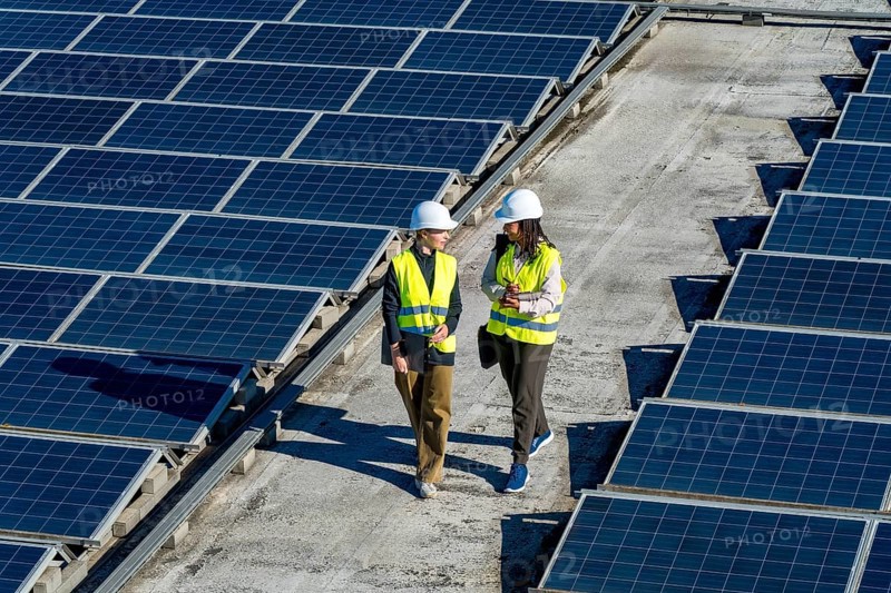Two women engineers wearing hard hats and high visibility vests walking and discussing work on a commercial rooftop with numerous solar panels. Symbolizing renewable energy and sustainable technology