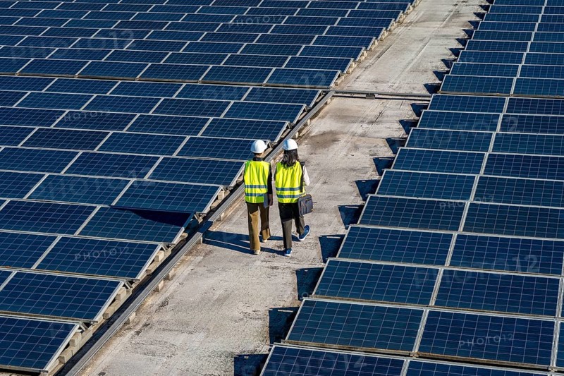 Two engineers, wearing hard hats and safety vests, walking between rows of solar panels on a rooftop, symbolizing renewable energy and sustainable power generation