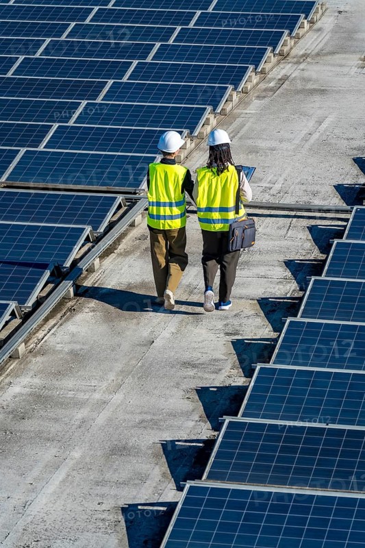 Two renewable energy engineers, wearing hard hats and reflective vests, walking along a rooftop solar power installation, performing maintenance and ensuring sustainable clean energy production