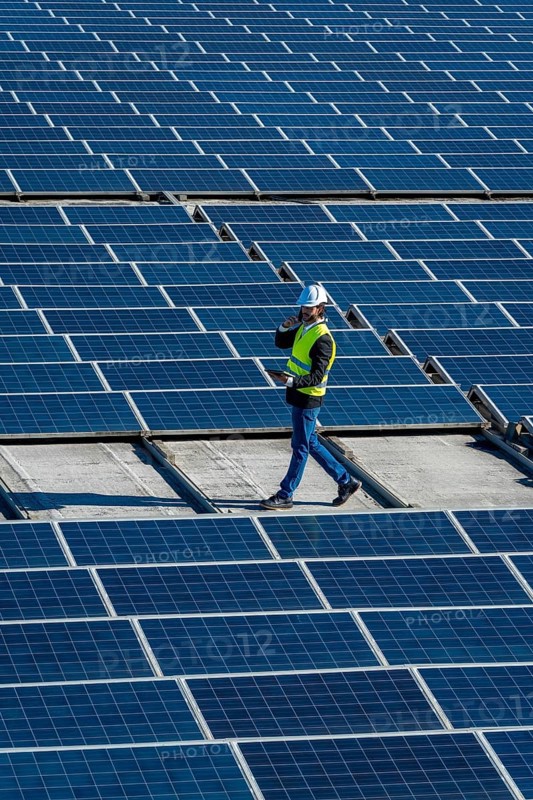 Engineer wearing safety helmet and vest, holding digital tablet and talking on phone while walking among rows of blue solar photovoltaic panels on a rooftop