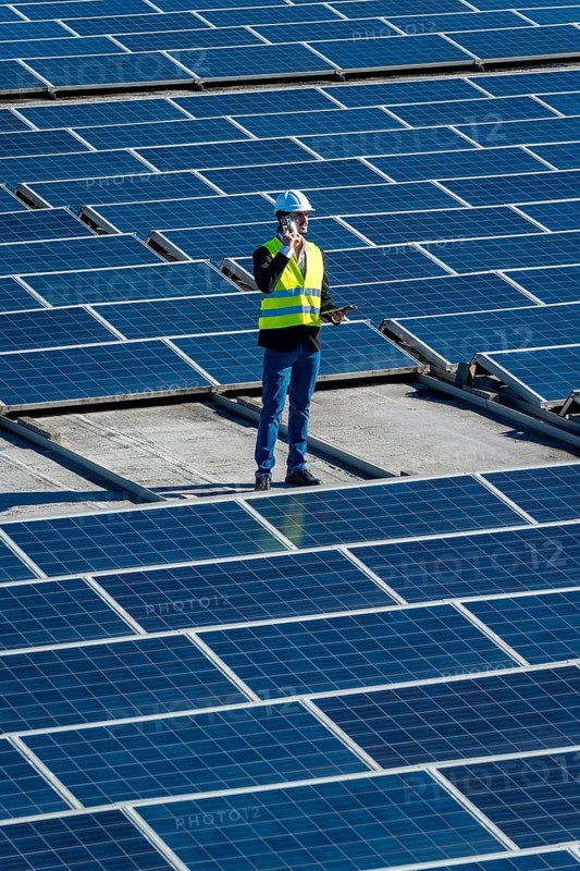 Engineer in hard hat and safety vest on commercial rooftop amid solar panels, using mobile phone while inspecting and maintaining photovoltaic array for clean, sustainable energy projects
