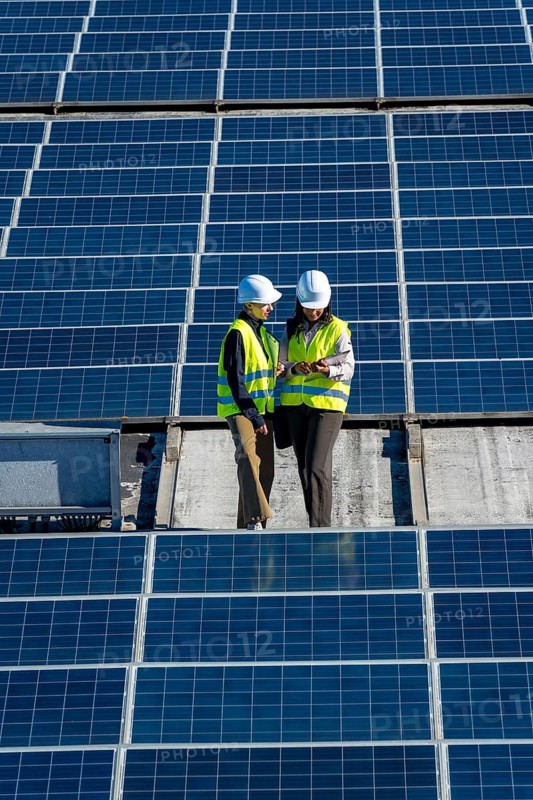 Female engineers wearing hard hats and high visibility vests discussing data on a digital tablet while standing on a rooftop solar panel installation, representing teamwork and renewable energy