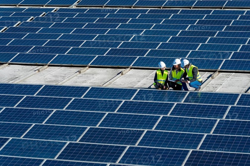 Team of diverse engineers wearing white hard hats and high visibility vests inspecting an array of solar panels, collaborating on sustainable energy generation and technological innovation