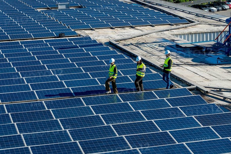Engineers and maintenance workers are walking across an expansive array of solar panels installed on a commercial building roof, performing an inspection of the renewable energy system