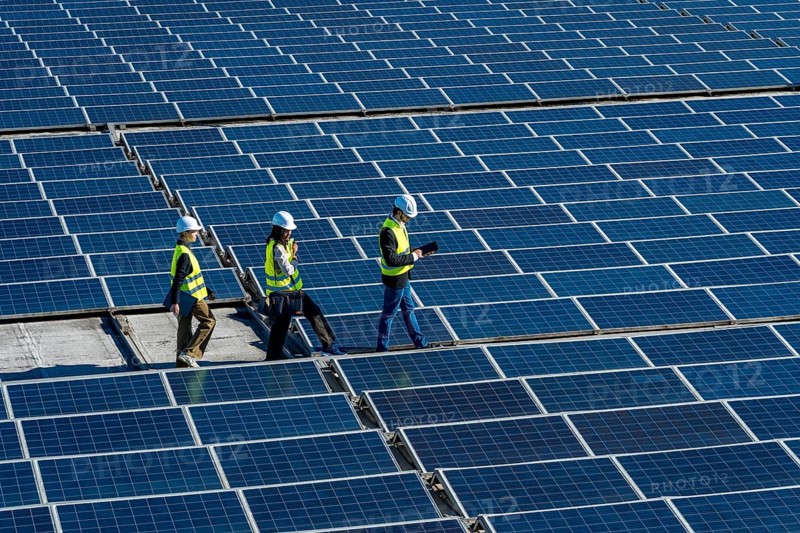 Engineers wearing safety helmets and vests are walking across a vast field of photovoltaic solar panels, examining the renewable energy installation and ensuring its optimal performance
