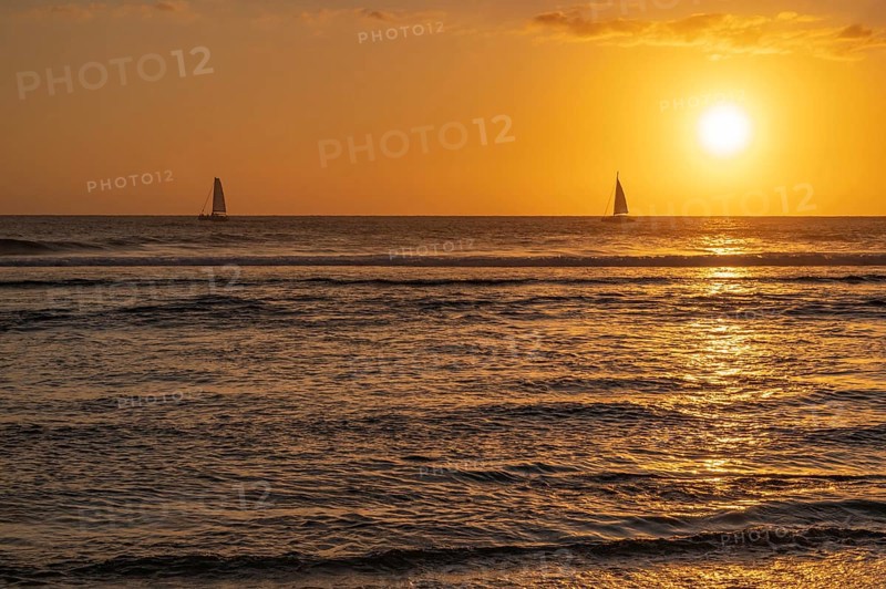 A warm sunset light bathes the sky over the Indian Ocean in orange and gold as two small sailboats cruise on the dark waves in the sea, Saint-Gilles, La Reunion, France