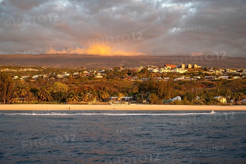 The bright sandy beach of the Saint-Gilles lagoon with bathers merges into a coastal landscape with palm trees and white houses, while the mountains are illuminated by warm evening light in the background and a jet ski ride through the sea, Saint-Gilles, La Reunion, France