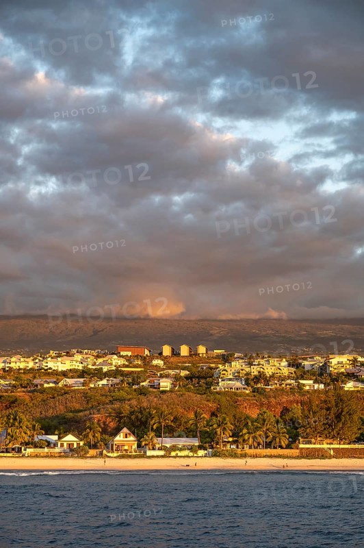 The bright sandy beach of the Saint-Gilles lagoon with bathers merges into a coastal landscape with palm trees and white houses, while the mountains are illuminated by warm evening light in the background, Saint-Gilles, La Reunion, France