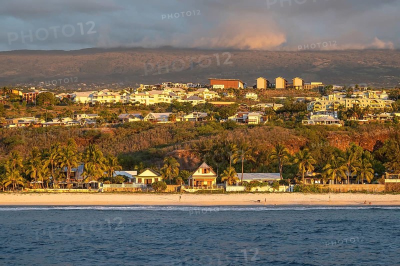 The bright sandy beach of the Saint-Gilles lagoon with bathers merges into a coastal landscape with palm trees and white houses, while the mountains are illuminated by warm evening light in the background, Saint-Gilles, La Reunion, France
