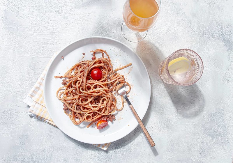 A plate with spaghetti topped with tomato sauce and cherry tomatoes sits on a light table. A glass of cool drink accompanies the meal, creating a relaxed dining atmosphere