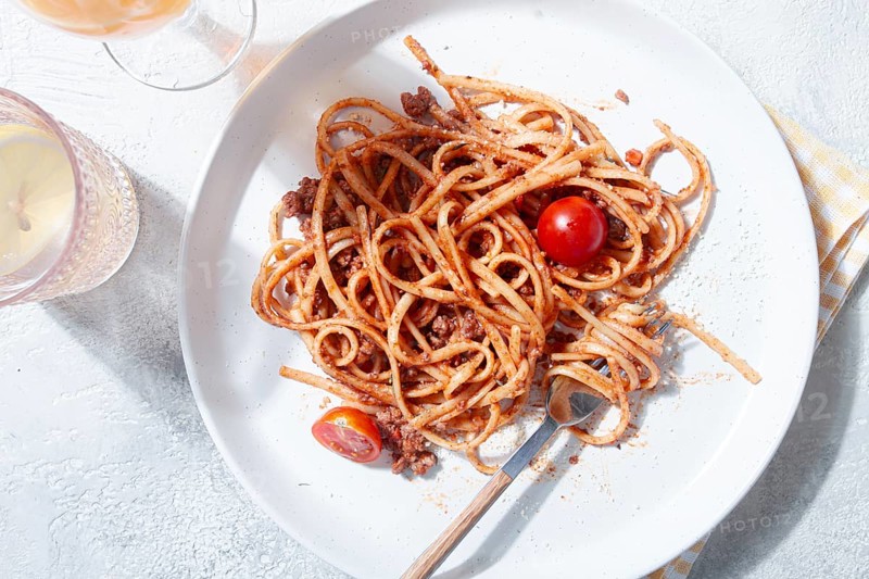 A plate holds freshly cooked spaghetti topped with flavorful meat sauce and garnished with bright cherry tomatoes. A glass sits nearby, reflecting a relaxed dining atmosphere