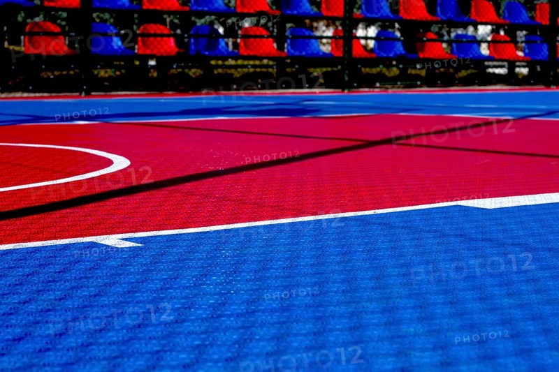 The basketball court features a striking red and blue design illuminated by bright sunlight. The court lines are clearly visible, creating a dynamic playing surface for sports activities