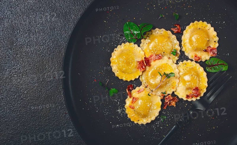 Fresh ravioli, with butter, spices and herbs, fried bacon, on a black plate, top view, dark background, no people