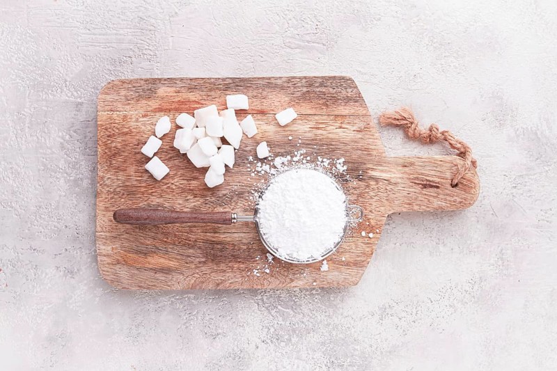 A strainer with powdered sugar, on a wooden cutting board, with pieces of white sugar