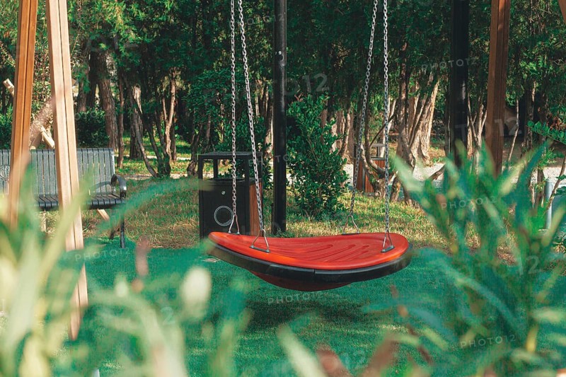 A vibrant red swing hangs suspended in a green park, surrounded by tall trees and wooden benches. The area appears peaceful with sunlight filtering through the leaves