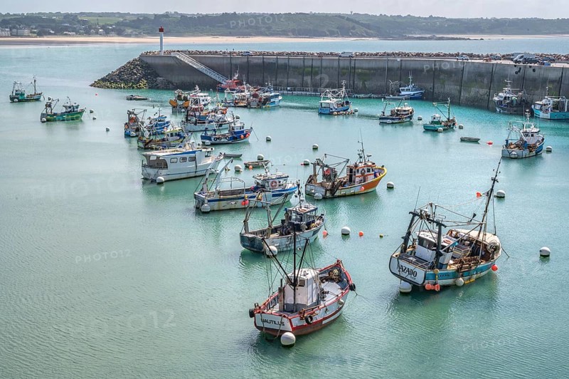 At low tide, numerous colorful, traditional fishing boats with netting equipment lie quietly moored in the turquoise water of the harbor basin, whose stone quay wall with the red and white lighthouse marks the harbor entrance in the background, Erquy, Saint-Brieuc Bay, Côtes-d'Armor, Brittany region, France