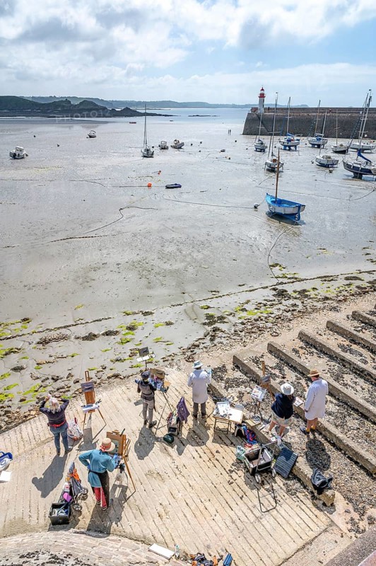 A group of plein-air amateur painters and artists stand with easels on a concrete-like ramp and looks at the large, muddy sandy beach, on which numerous sailing and fishing boats dry at low tide and in the background the pier with the small, red and white lighthouse marks the harbor entrance, Erquy, Saint-Brieuc Bay, Côtes-d'Armor region, France