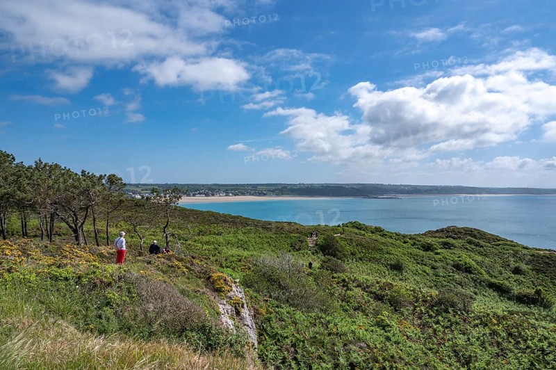 An elevated panorama shows the vast landscape of Cap d'Erquy covered with pine, broom and low scrub, from which a hiking couple looks across the extensive bay with the village's sandy beach and the emerald green sea, Erquy, Cap d'Erquy, Côtes-d'Armor department, Brittany region, France