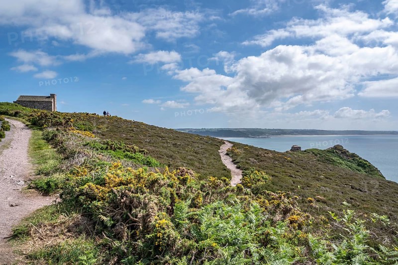 An unpaved coastal path, the Sentier Littoral GR 34 long-distance hiking trail, snakes across the cliff covered with yellow broom and heather, on whose hill the historic stone building of the Corps de Garde des Trois Pierres looks across the bay and the vast sea, here the Atlantic, Erquy, Cap d'Erquy, department of Côtes-d'Armor, Brittany region, France