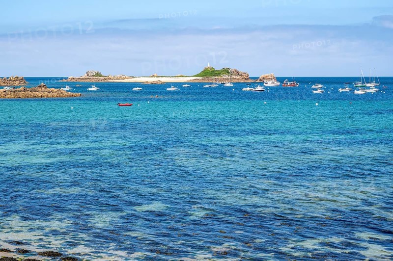 A large rock of pink granite shaped by wind and weather dominates the right foreground, while in the background the bright turquoise-blue water of the Atlantic is crossed by numerous sailboats and the small, island-like headland with the Saint-Gildas lighthouse, Port Blanc, Penvenan, Cotes-d'Armor department, Brittany region, France
