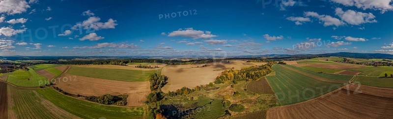 A large, open landscape shows green and brown fields with scattered trees. The sky is clear and sunny, clouds are passing by. Železná hurka, Czech Republic