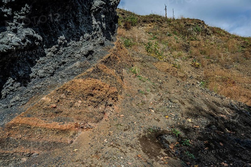 The scene shows different layers of earth and rock formations in a rural area on a clear day. Dry grass and small plants are visible. Železná hurka, Czech Republic