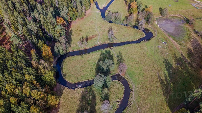 A clear river snakes through a green meadow, surrounded by colorful autumn trees. The picture shows the tranquil beauty of nature during autumn. Falkenberg, Upper Palatinate