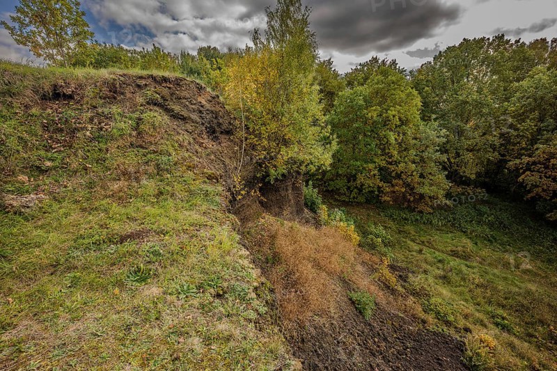 The gentle slope of an extinct volcano shows a variety of trees with colorful autumn leaves. The surrounding area is quiet and offers a beautiful view of green grass and wooded areas. Železná hurka, Czech Republic