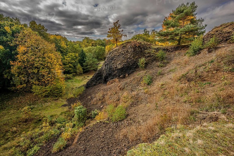 The scene shows a varied autumn landscape with colorful trees. The sky is cloudy and makes the colors of nature appear vivid. The slope of the extinct volcano is clearly formed. Železná hurka, Czech Republic