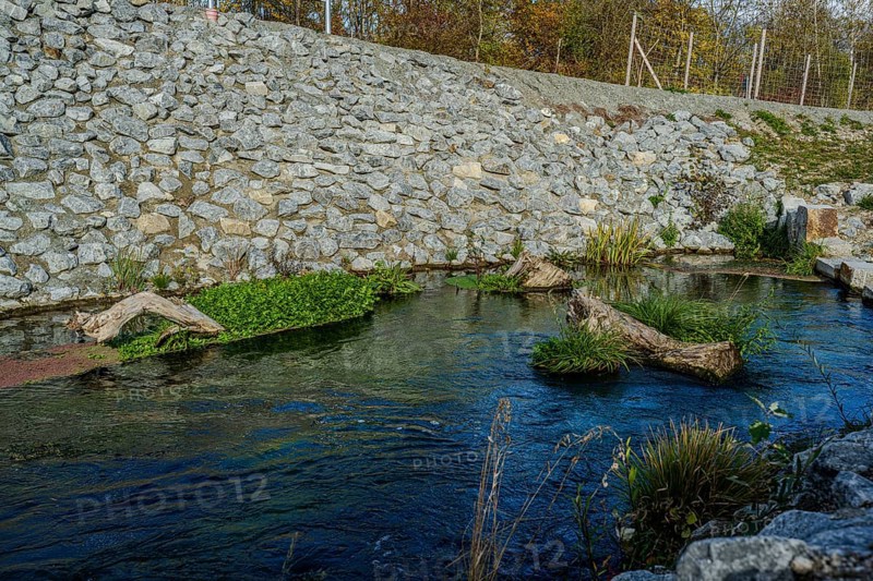 A calm stream with clear water flows through an area lined with stones. Green plants and pieces of wood lie in the water. The colors change due to the soft autumn light. Landau an der Isar, Lower Bavaria