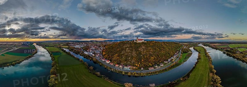 The Bogenberg with the pilgrimage church of the Assumption of the Virgin rises above the quiet Danube. The surrounding area shows green fields and the clouds reflect the light of the setting sun. Bogen, Lower Bavaria