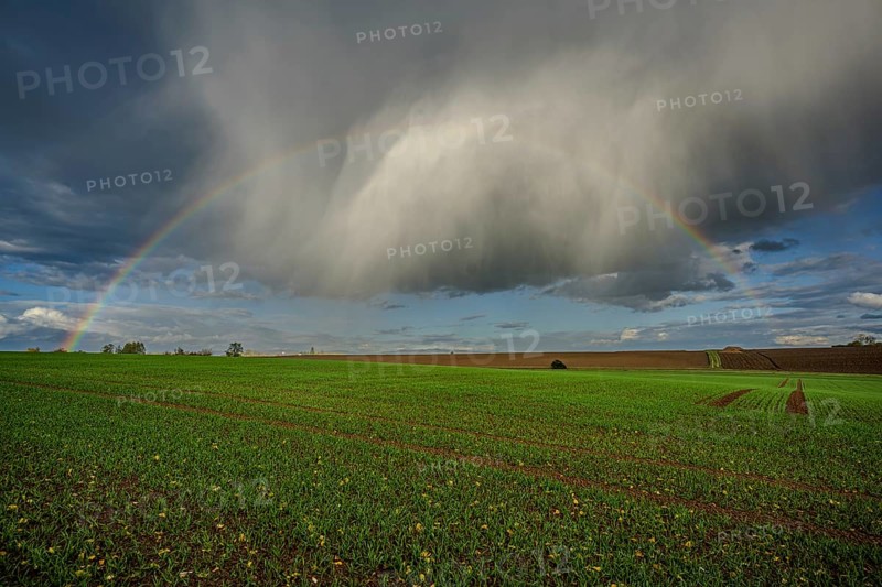A rainbow stretches over a lush green field, while the clouds in the background gather after the rain. The rural environment gives off a peaceful feeling. Landau an der Isar, Lower Bavaria