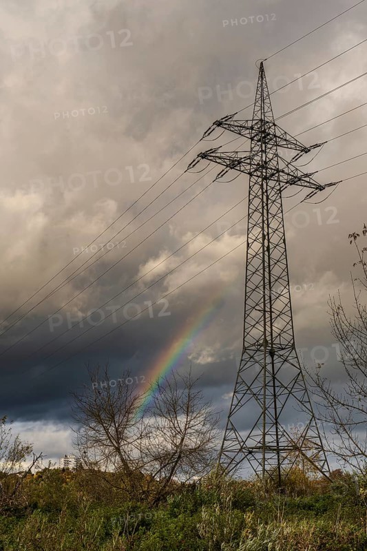 There is a high-voltage pylon in a rural area. Behind the mast, a rainbow appears in the sky. It is cloudy and looks like a thunderstorm. Bogen, Lower Bavaria