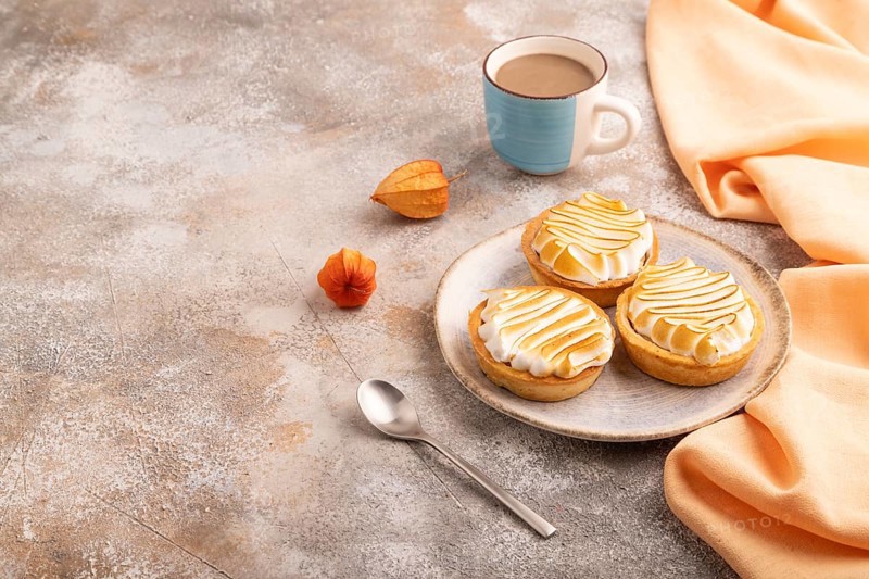 French lemon tart with meringue on brown concrete background, cup of coffee, orange linen textile, side view, copy space