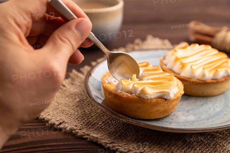 French lemon tart with meringue with hand on brown wooden background, cup of coffee, linen textile, side view, close up, selective focus