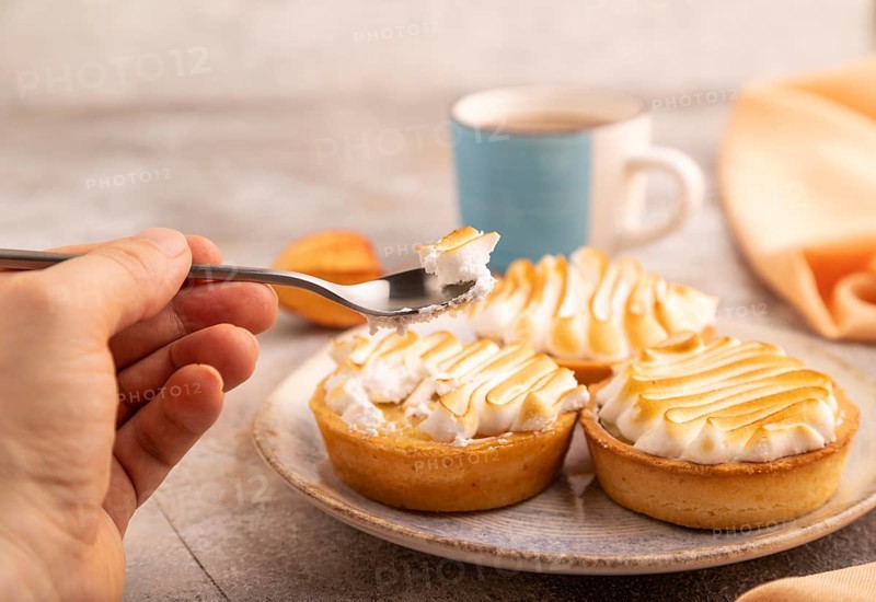 French lemon tart with meringue with hand on brown concrete background, cup of coffee, orange linen textile, side view, close up, selective focus