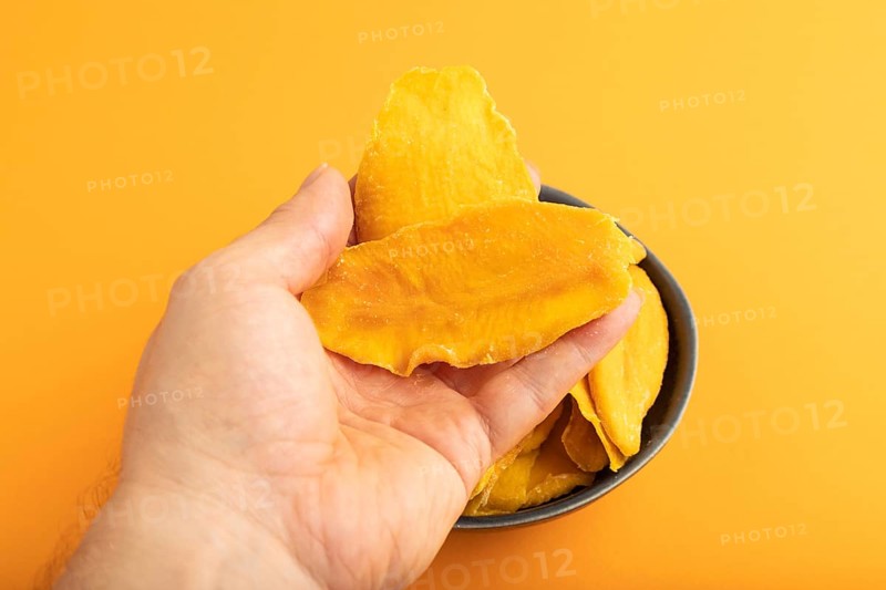 Dried Mango in blue ceramic bowl with hand on orange pastel paper background. Side view, close up. healthy food, minimalism