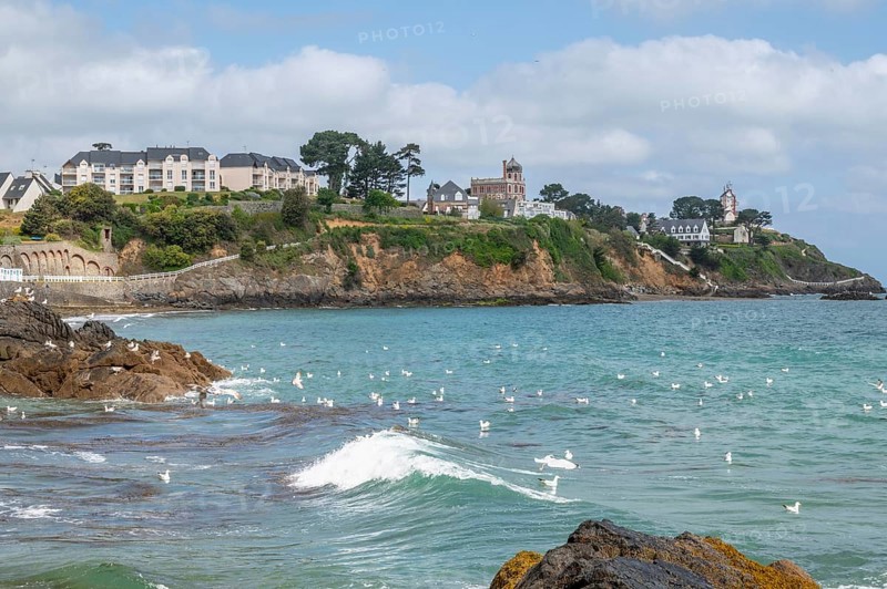 The rocky bay of the Plage de la Comtesse with a little wave wave in the turquoise water, which is bordered in the background by a headland built with villas and the offshore Ile de la Comtesse as seagulls fly across the sea, Saint-Quay-Portrieux, Saint-Brieuc Bay, Cotes-d-Armor, Brittany, France