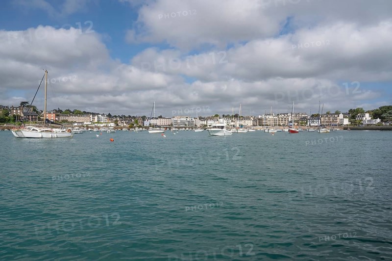 A series of small sailboats, probably optimists, with red and white ringed sails, move in formation parallel to the coast across the slightly curled turquoise water, behind which a beacon can be seen on a rocky headland, Plerin, Saint-Brieuc Bay, Cotes-d-Armor, Brittany, France