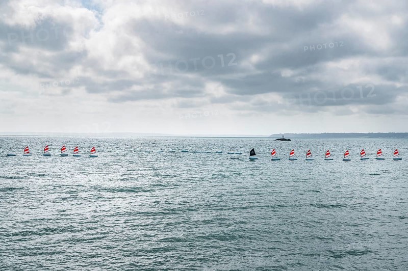 A series of small sailboats, probably optimists, with red and white ringed sails, move in formation parallel to the coast across the slightly curled turquoise water, behind which a beacon can be seen on a rocky headland, Plerin, Saint-Brieuc Bay, Cotes-d-Armor, Brittany region, France