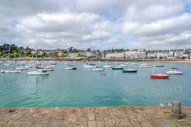 The view from a stone harbor wall shows numerous sailing and motor boats on the turquoise water of the harbor basin, behind which the dense, multi-storey development of the seaside resort stretches along the coastline, Saint-Quay-Portrieux, Cotes-d'Armor region, France