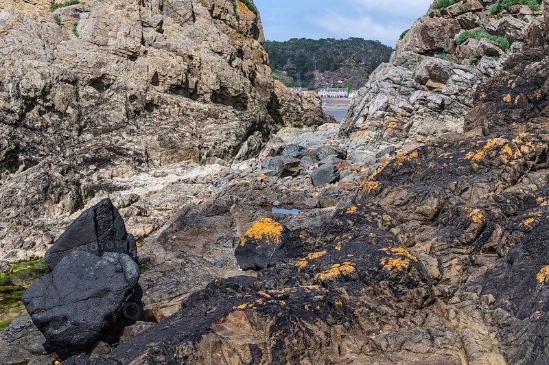 A rugged rock formation, dark and yellow-orange covered with lichens, which forms a narrow opening through which the wide sandy beach and the buildings of the town of Erquy can be seen in the distance, Erquy, Saint-Brieuc Bay, Côtes-d'Armor, Brittany region, France