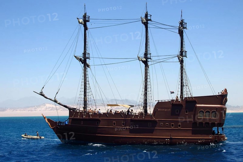The luxury pirate yacht Black Pearl, with a group of tourists on board, enjoys a cruise through the vast tropical expanses of the Red Sea. The vessel is located near the seaport of the resort town of Sharm el-Sheikh, where tourists and travelers depart for boat excursions. The desert and mountains of the Sinai Peninsula are visible in the background. South Sinai, Sharm el-Sheikh, Egypt