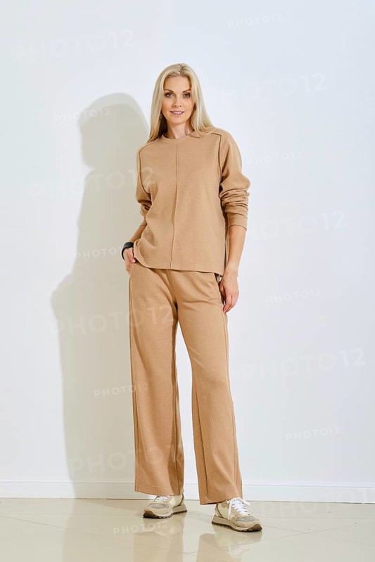 A woman stands confidently in a casual beige outfit featuring a sweater and loose pants. She poses against a plain white wall, showcasing a modern and relaxed style. Natural light brightens the room