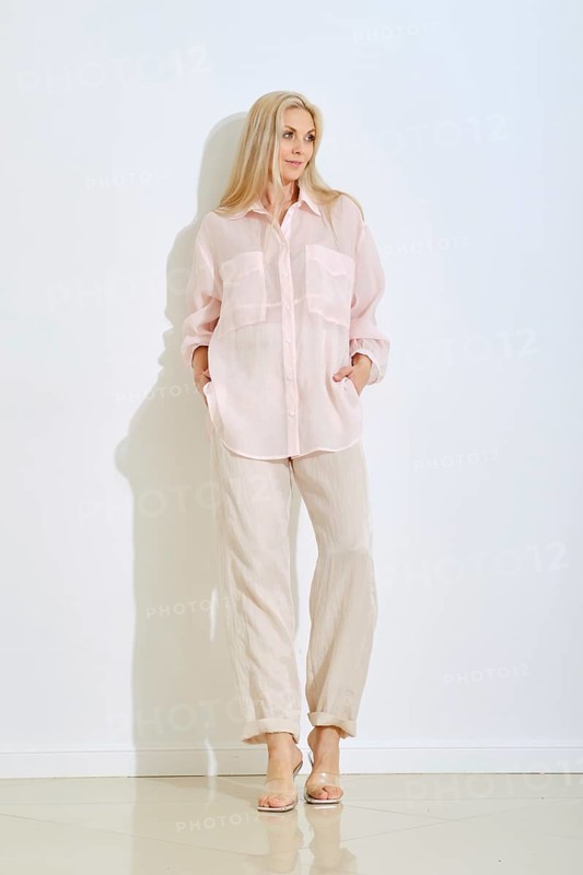 A model stands confidently in a light pink button-up shirt and beige pants. She poses with hands in pockets, showcasing her relaxed style in a minimalist indoor setting with a white wall