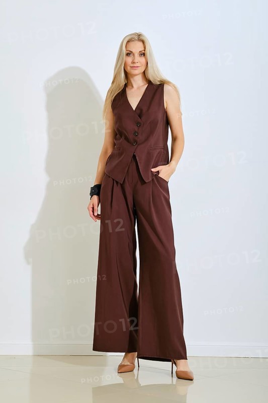 A woman stands elegantly in a chic brown tailored outfit that includes a fitted vest and wide-leg trousers. Her long hair falls gracefully as she strikes a confident pose