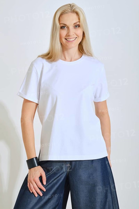 A woman stands confidently, wearing a plain white t-shirt paired with loose denim pants. She smiles warmly, with soft lighting highlighting her features in a bright indoor environment